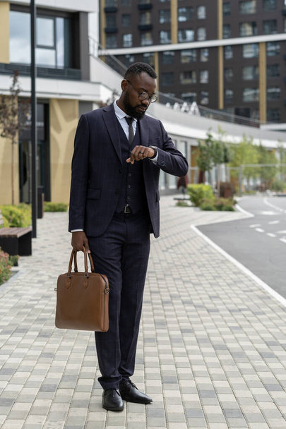 Elegant businessman with handbag looking at wristwatch while standing on road - Foto, imagen