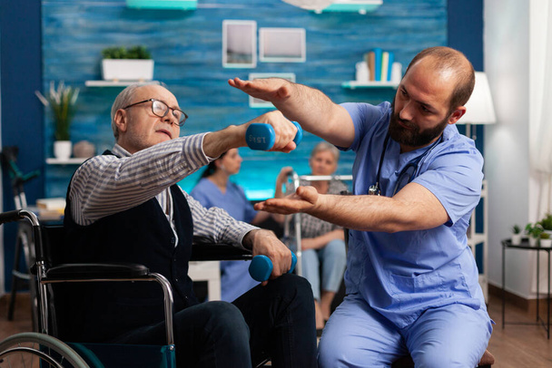 Врач-практикант помогает пожилому мужчине в инвалидном кресле выполнять физиотерапевтические упражнения - Фото, изображение