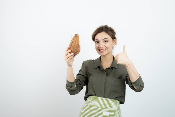 Picture of young woman in apron showing thumb up and holding a coconut . High quality photo - Foto, Imagem