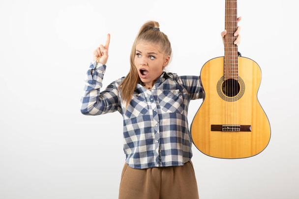 Φωτογραφία της νεαρής ξανθιάς που δείχνει πάνω και κρατάει ακουστική κιθάρα. Υψηλής ποιότητας φωτογραφία - Φωτογραφία, εικόνα