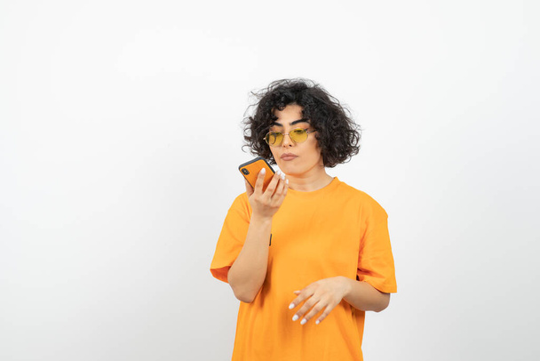 donna in occhiali e t-shirt arancione in posa con smartphone in studio - Foto, immagini