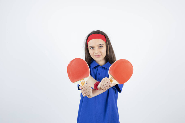 niña en camiseta azul, pulsera roja y diadema posando con raquetas de tenis - Foto, Imagen