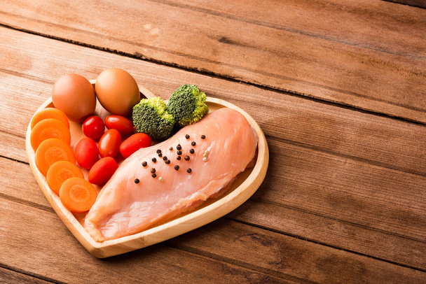 Top view of Raw chicken breasts fillets no boneless with spices rosemary, carrot, and eggs in heart plate wood on wooden background, Healthy food day concept - Фото, зображення