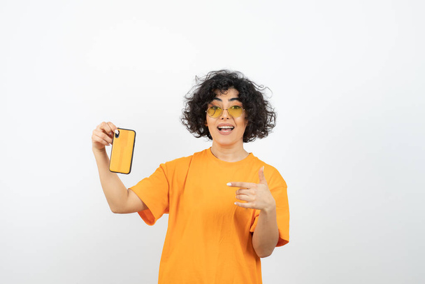 woman in glasses  and orange t-shirt posing with smartphone in studio - Foto, Imagen