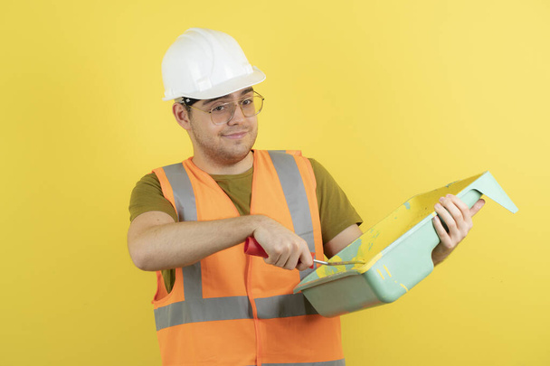 Jeune ouvrier de la construction en hardhat prenant de la peinture jaune avec rouleau de peinture. Photo de haute qualité - Photo, image