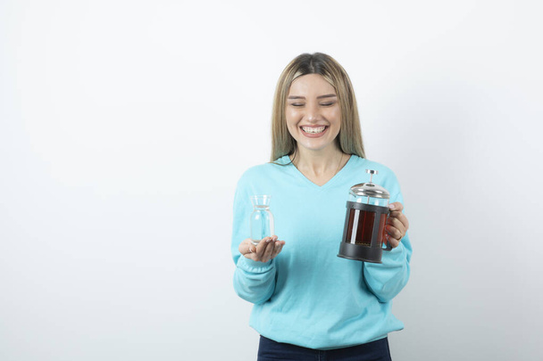 Positive cheerful girl holding teapot with a glass cup against white wall. High quality photo - Фото, изображение