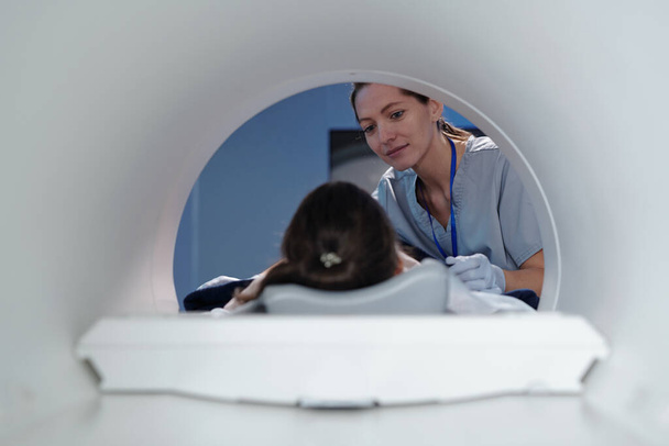 Jovem clínico feminino olhando para a menina deitada na mesa de ressonância magnética - Foto, Imagem
