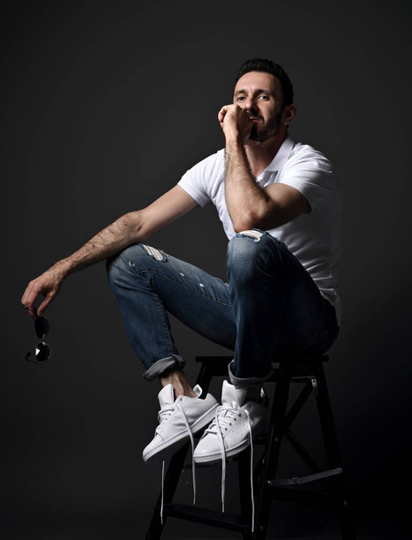 Thoughtful unshaved man in white polo shirt, modern torn jeans and sneakers sits on high chair, stepladder - Foto, afbeelding