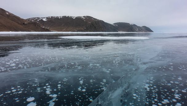La superficie lisa y brillante de un lago congelado. Las grietas penetran profundamente en el hielo. Burbujas de gas metano son visibles. Una cordillera contra el cielo. Baikal. - Foto, imagen