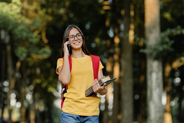 Красива студентка в окулярах і червоний рюкзак ходить по кампусу і розмовляє по телефону після навчання
 - Фото, зображення