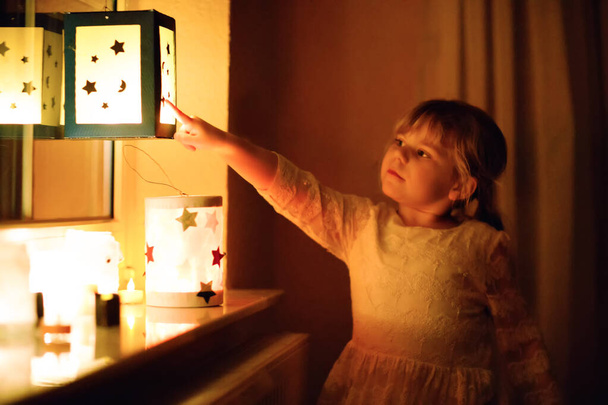 Klein meisje zit bij het raam met zelfgemaakte handgemaakte lantaarns met kaarsen voor St. Martin processie. Peuterkind kijkt naar gloeiende lantaarn. Duitse traditie Martinsumzug. Huisdecoratie - Foto, afbeelding