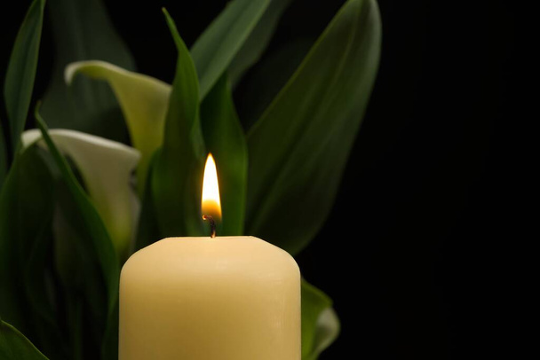 Close up of candle burning flame in the darkness and arum lilies illuminated by the candlelight alongside in a conceptual image - Photo, Image