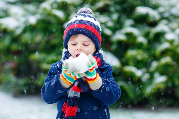 Cute little funny dziecko w kolorowe ubrania mody zimowej zabawy i zabawy ze śniegiem, na zewnątrz podczas opadów śniegu. Aktywny wypoczynek na świeżym powietrzu z dziećmi. Chłopiec i maluch łapie płatki śniegu. - Zdjęcie, obraz