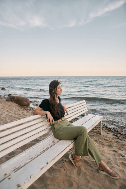 Όμορφη μελαχρινή γυναίκα κοιτάζει μακριά στον ορίζοντα της θάλασσας. Τα κορίτσια κάθονται σε λευκό παγκάκι στην παραλία. Έννοια της ισορροπίας και της ευημερίας της εργασίας και της ζωής. - Φωτογραφία, εικόνα