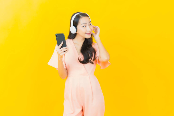 Portrét krásná mladá asijská žena se sluchátkem a chytrý telefon pro poslech hudby na žlutém pozadí - Fotografie, Obrázek