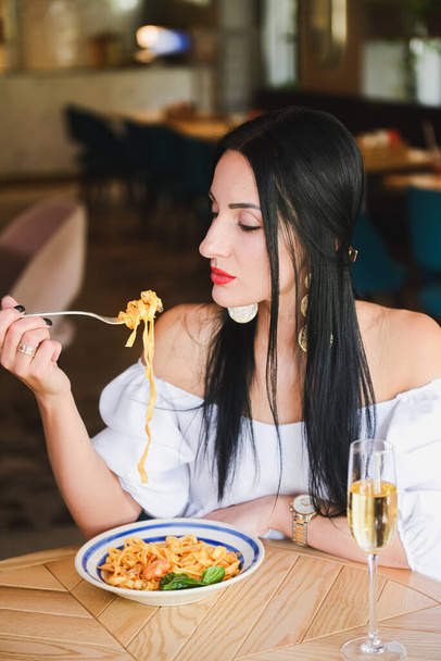 Gorgeous young brunette woman having Italian food. Beautiful woman inside Italian restaurant eating traditional Italian pasta spaghetti. Alone in restaurant, social distancing. Fashion shooting. - Фото, изображение
