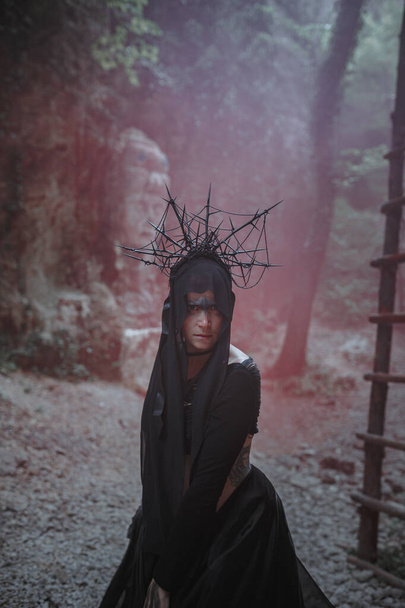 魔女の若い女性は黒のドレスに立って、暗い霧の森の中で彼女の頭の上に冠。ハロウィンコスチュームのためのダークパーティー. - 写真・画像