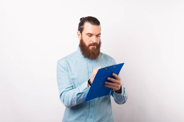 Serious bearded man is writing something on a paper holder board. - Фото, изображение