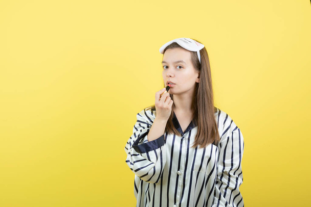  Beautiful young woman standing and putting lipstick against yellow wall . High quality photo - Fotó, kép
