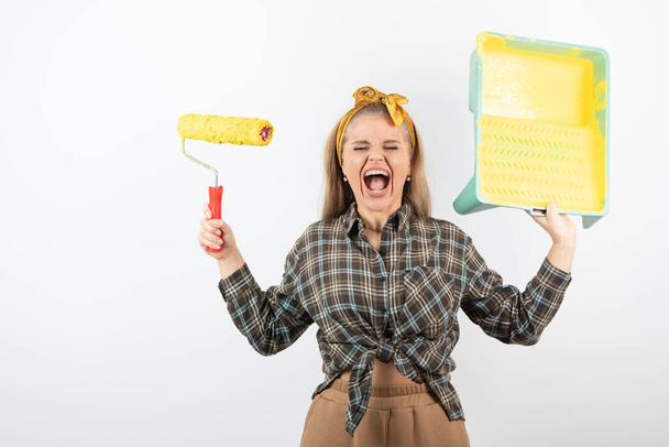 Young blonde woman holding roller tray with yellow paint roller over a white wall. High quality photo - Photo, Image