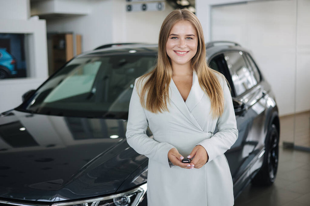 Portrait of beautiful and young business woman in car showroom. Female hold keys from her new car - 写真・画像