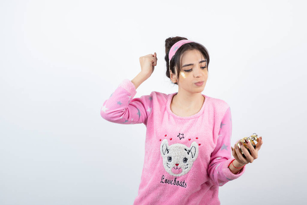 Młoda dziewczyna w różowej piżamie patrząc na czas na budziku nad szarą ścianą. Wysokiej jakości zdjęcie - Zdjęcie, obraz