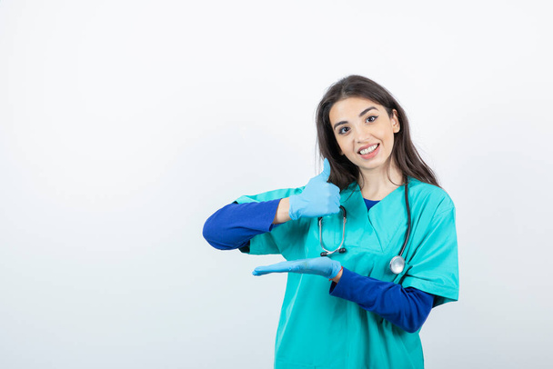 Retrato de jovem enfermeira com estetoscópio em luvas médicas mostrando o polegar para cima. Foto de alta qualidade - Foto, Imagem