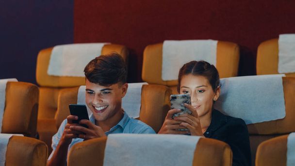 Atractiva joven pareja caucásica alegre usando un teléfono inteligente mientras ve el cine en el cine. Concepto de entretenimiento de estilo de vida. - Foto, Imagen