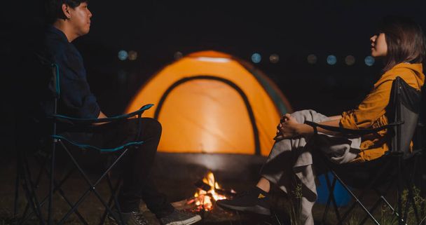 Young asia couple backpackers enjoying in camping at night near campfire on beach. Male and female traveler relaxing and talking at campsite. Outdoor activity, adventure travel, or holiday vacation. - Photo, Image
