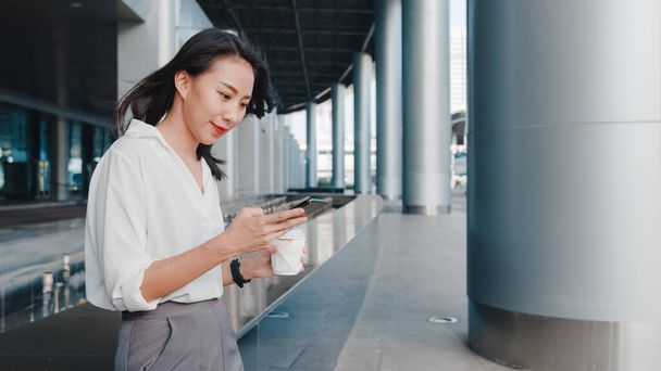Successful young Asia businesswoman in fashion office clothes holding disposable paper cup of hot drink and using smart phone while walking outdoors in urban modern city. Business on the go concept. - Fotoğraf, Görsel