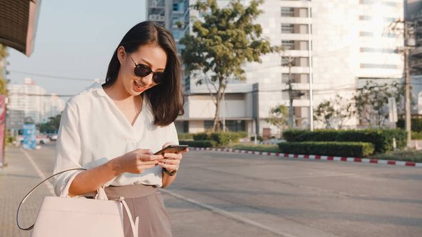 Successful young Asia businesswoman in fashion office clothes hailing on road catching taxi and holding smart phone while standing outdoors in urban modern city. Business on the go concept. - Photo, Image