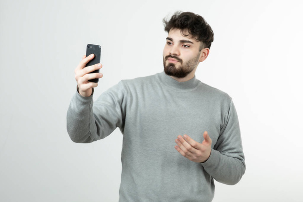 Portrait d'un gars attrayant tenant un téléphone intelligent contre un mur blanc. Photo de haute qualité - Photo, image