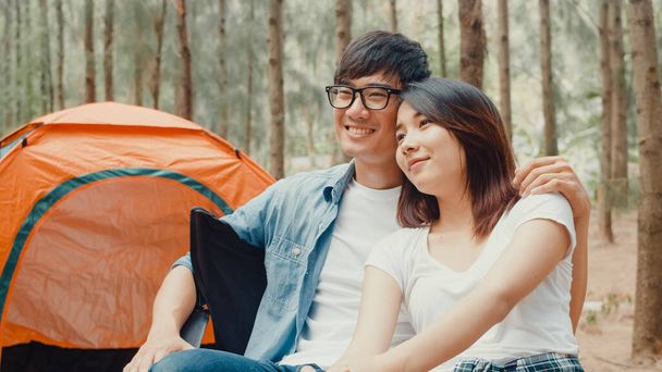 若いアジアのキャンパーカップルの椅子にテントで森の中に座っている。男性と女性の旅行者は、キャンプ場で夏の日にリラックスして話をします。野外活動、冒険旅行、または休暇. - 写真・画像