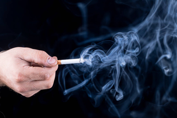 Černé pozadí. v ruce muže s drsnou kůží drží cigaretu. Vychází z ní modrý kouř. close-up - Fotografie, Obrázek