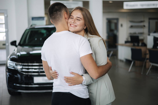 Счастливая женщина обнимает своего мужа, покупая машину в автосалоне. Мужчина и женщина покупают новую машину - Фото, изображение