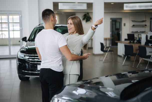 幸せな女性ははるかに車のショールームで車を購入する彼の夫を抱擁。男と女が新車を買う - 写真・画像