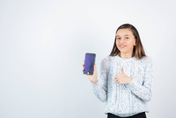 Mädchen im Pullover posiert mit Smartphone auf weißem Hintergrund - Foto, Bild