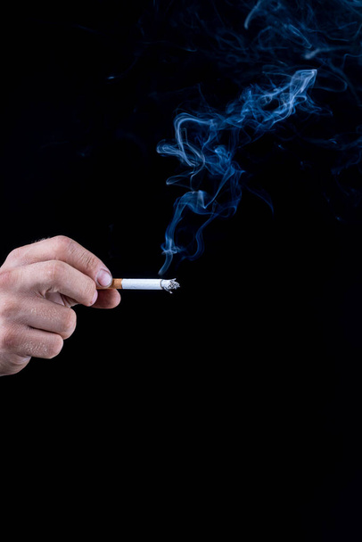 Fond noir. la main d'un homme à la peau rugueuse tient une cigarette. de la fumée bleue en sort. gros plan - Photo, image