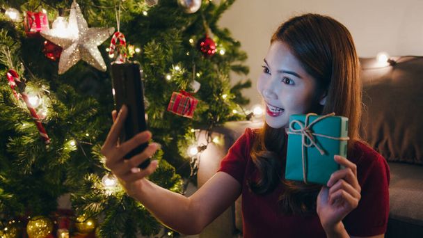Młoda Azja kobieta za pomocą smartfona rozmowy wideo z parą z X 'Mas pudełko prezentów, choinka ozdobiona ozdobą w salonie w domu. Świąteczna noc i święto Nowego Roku. - Zdjęcie, obraz