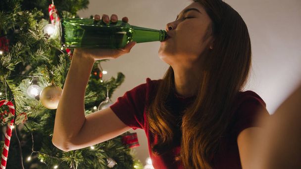 幸せな夜のパーティービデオ通話を楽しんでいる若いアジアの女性がビールを飲むカップル、自宅のリビングルームで装飾が施されたクリスマスツリーと話をします。クリスマスの夜と新年の休日の祭り. - 写真・画像