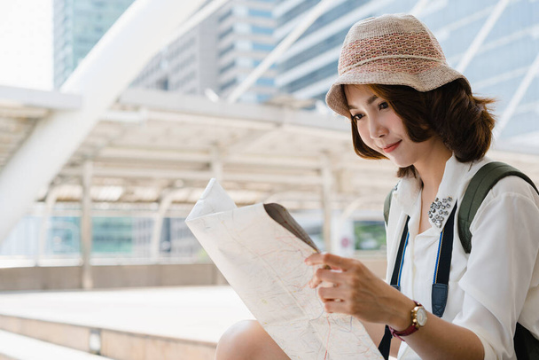 Happy Asian woman traveler tourist wanderer with trendy look searching direction on location map while traveling abroad in summer. Young female traveler with backpack and with map on street in city. - Photo, Image
