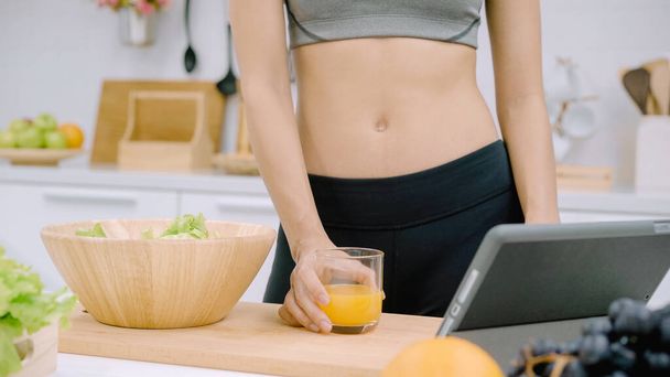 Young Asian woman making salad healthy food while using tablet for looking recipe and drinking orange juice in the kitchen, beautiful female preparing salad for fit body at home. Healthy food concept. - Foto, imagen