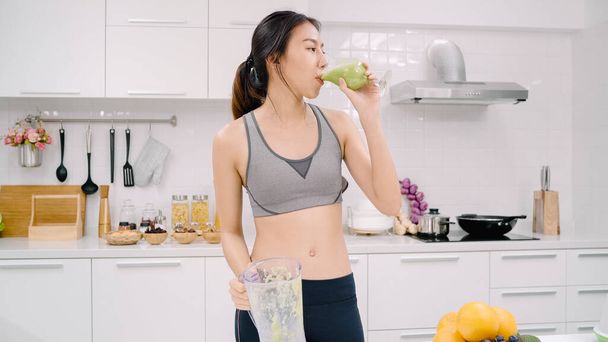 Σπορ ασιατική γυναίκα πίνοντας χυμό μήλου στην κουζίνα, όμορφη γυναίκα σε αθλητικά ρούχα Χρησιμοποιήστε πολλά βιολογικά φρούτα διατροφή παραγωγή χυμού Μήλου από τον εαυτό της στο σπίτι. Έννοια της υγιεινής διατροφής. - Φωτογραφία, εικόνα
