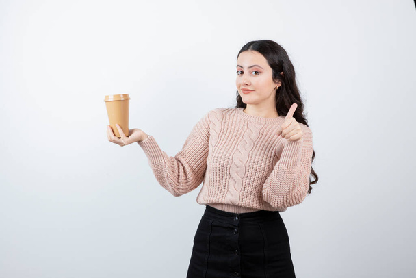 Молода брюнетка показує великий палець і тримає чашку гарячої кави. Високоякісна фотографія
 - Фото, зображення