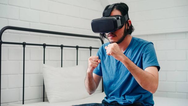 Young Asian man using glasses virtual reality simulator playing boxing game in bedroom, Japanese male feeling happy using relax time lying on bed at home. Teen play games relax at home concept. - Fotó, kép