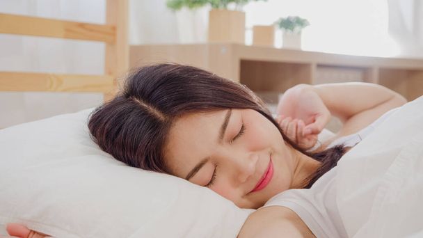Kaunis houkutteleva nuori aasialainen nainen herää, hymyilee ja venyttelee käsiään sängyssä makuuhuoneessa. Nuori aasialainen nainen käyttää rentoutumisaikaa kotona. Elämäntapa naiset rentoutua kotona käsite. - Valokuva, kuva