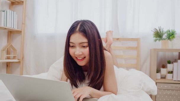 Młoda firma freelance Asian kobieta pracuje na laptop sprawdzanie mediów społecznościowych leżąc na łóżku, gdy relaks w sypialni w domu. Kobiety Lifestyle w koncepcji domu. - Zdjęcie, obraz