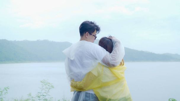 Young Asian sweet couple feeling happy using romantic time playing rain while wearing raincoat standing near lake. Lifestyle couple enjoy and relax in rainy day. - Photo, Image