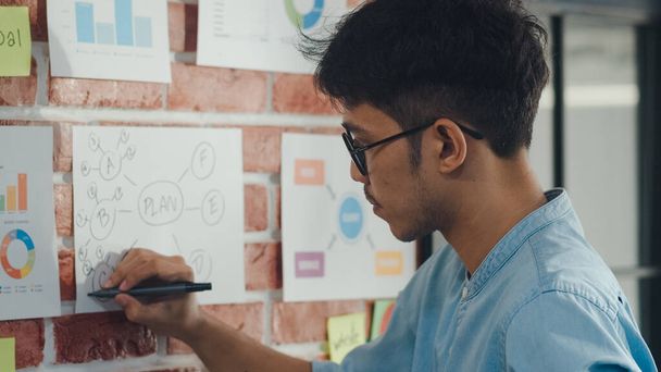 asiatische kreative Mann zeichnen Arbeitsplan auf Pappe. junge professionelle Unternehmer männlich denken und schreiben Informationen Erinnerung auf Papier auf Ziegel, Geschäftslage, Start-up in Loft-Office-Konzept. - Foto, Bild