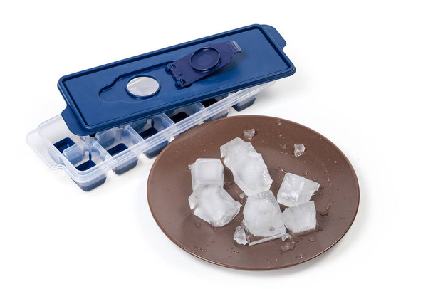 Cubos de hielo en plato marrón contra el molde para hacer cubitos de hielo domésticos con tapa removida sobre un fondo blanco - Foto, imagen